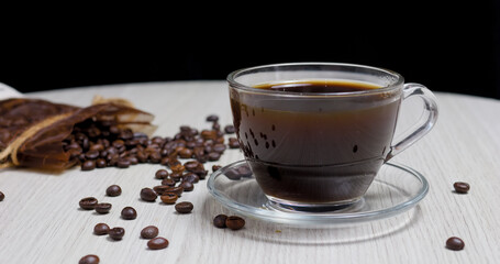 Steaming cup of black coffee sits on a glass saucer on a light wooden table, surrounded by roasted coffee beans spilling from a rustic burlap bag, creating a cozy and inviting atmosphere