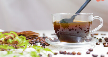 Stirring black coffee in a clear glass cup resting on a saucer, surrounded by scattered coffee beans and a vibrant green plant, creating a cozy atmosphere on the table