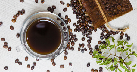 Glass cup of coffee sits on a white table, surrounded by roasted coffee beans spilling from a burlap bag and a fresh green sprig, creating a cozy and aromatic scene
