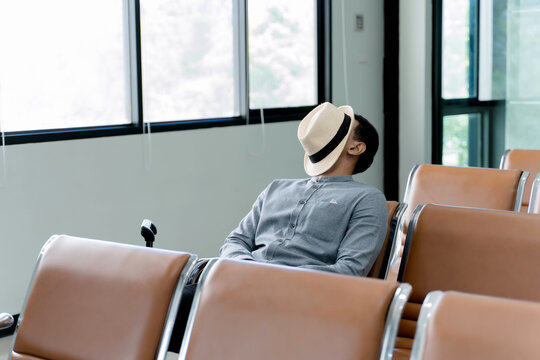Young man sleeping alone in airport waiting area, relaxing on chair with hat covering face, resting during layover or travel delay in empty terminal lounge