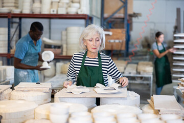 Senior woman ceramist in apron working in pottery workshop, looking at new crafted decorative plates.