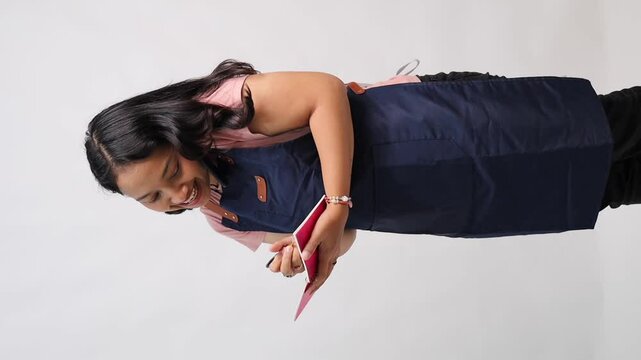 Smiling woman wearing a navy apron and pink shirt, looking at a red notebook or greeting card with joy and curiosity