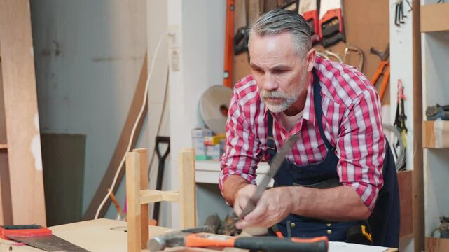Senior carpenter uses a ruler and set square to measure and assemble a wooden structure with precise alignment, focusing on craftsmanship. Workshop is organized with tools and materials
