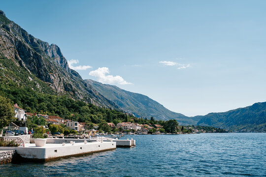 White pier on the shore of the bay against the backdrop of ancient houses at the foot of the mountains. Dobrota, Montenegro