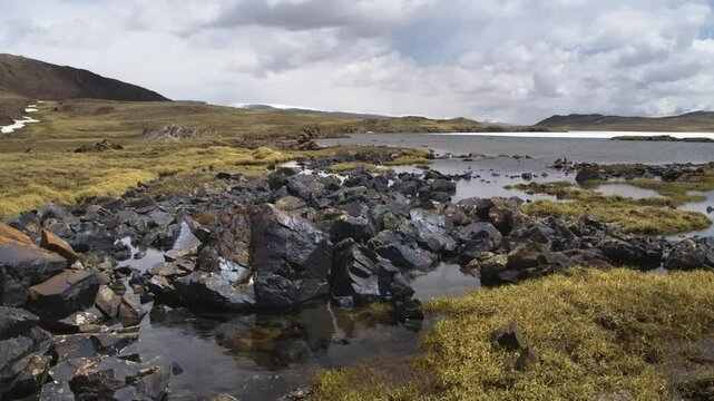 Footage depicts the stunning landscape of Arabel Plateau, Kyrgyzstan, showcasing a rocky shoreline with dark, volcanic-looking rocks and vegetation along a water body.