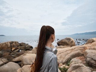 Young woman with long hair in gray hoodie stands by rocky beach, gazing at ocean, evoking emotions of contemplation and serenity during a cloudy day