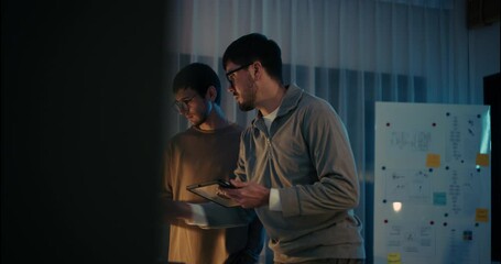 Two male developers reviewing project data on a tablet and smartphone in a tech office at night, with brainstorming board in the background. - Powered by Adobe