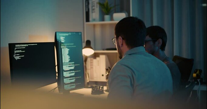 Two software developers working late at night, analyzing code on dual monitors in a modern tech workspace, symbolizing teamwork and productivity.