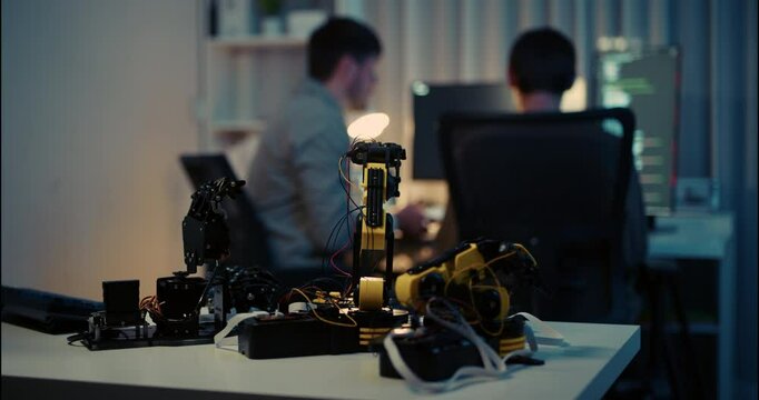 Robotic arms on a workspace desk, with two developers working in the background, symbolizing robotics, AI development, and coding for automation.