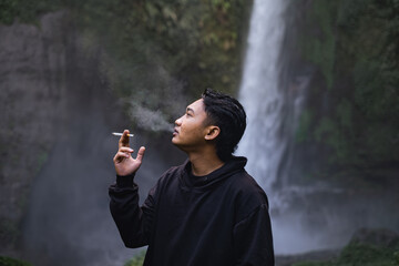 Southeast Asian man in black hoodie smoking a cigarette in the waterfall with bokeh background