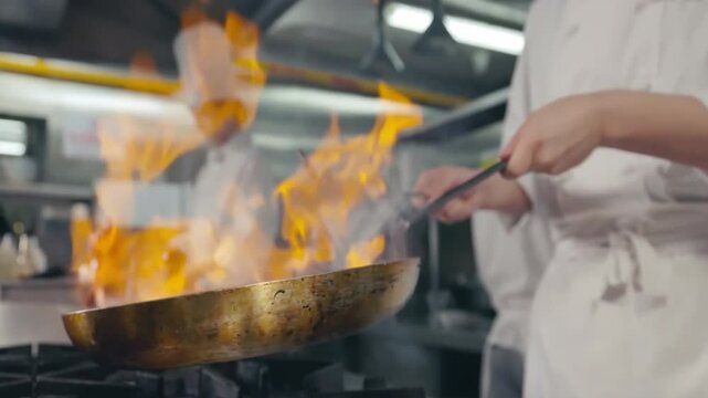 White uniform Professional Chef hold wok cooking vegetable stir fry in pan at restaurant kitchen. Flambler or cooking vegetables with spices on natural fire