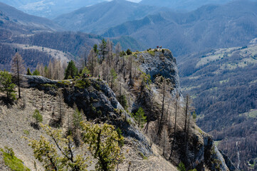 A scenic view of hikers on a rocky mountain ridge with expansive forested hills in the background under a clear blue sky.