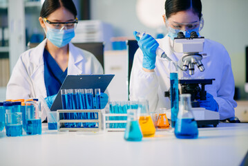 Fototapeta premium Two Asian female biotechnologists in lab coats conduct research, handle glassware, and test chemicals in a modern science lab