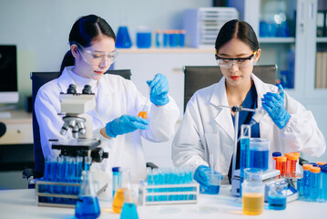 Fototapeta premium Two Asian female biotechnologists in lab coats conduct research, handle glassware, and test chemicals in a modern science lab