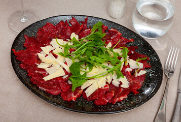 Thin slices of meat carpaccio, cheese and arugula on white plate served on table in restaurant. Traditional Italian dish
