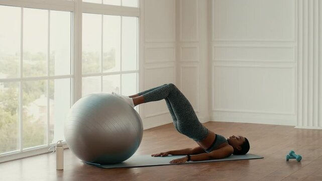 An African American woman engages in a workout session at home, using a stability ball for core strength training. The bright indoor space is designed for a healthy lifestyle.