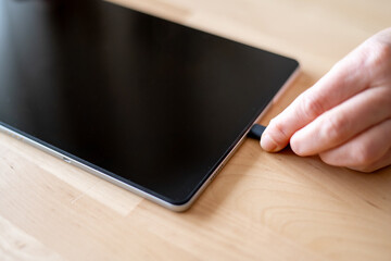 Hand inserting a usb cable into a tablet resting on a wooden table, preparing for charging or data transfer. Close-up detail highlighting the connection between technology and modern devices