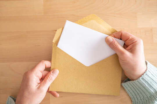 Woman's hands taking a blank white letter out of a brown envelope placed on a wooden table, offering copy space for customization and personalization