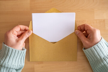 Woman's hands gently opening a brown envelope, revealing a blank white letter on a wooden table, ideal for adding personalized messages or creative designs