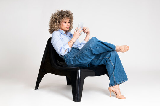 Fashionable woman with curly hair using smartphone in studio setting