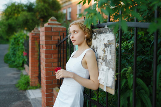 Young woman in a white dress with blonde hair leaning against a fence on a summer evening