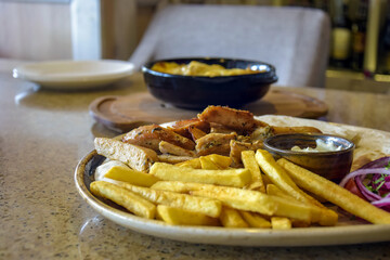 French fries with fried pork meat  in large ceramic dish, served with white sauce in separate saucer, sliced ​​blue onions, on marble table. Lunch in cafe. Tasty unhealthy food. No people. Copy space