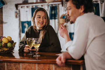A woman and a man enjoy friendly conversation with glasses of white wine at a kitchen counter, evoking a casual, warm, and inviting social setting.