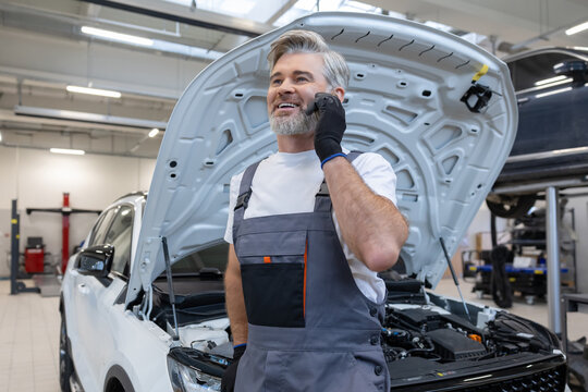 Male mechanic in garage holding smartphone talking about automobile repair with customer