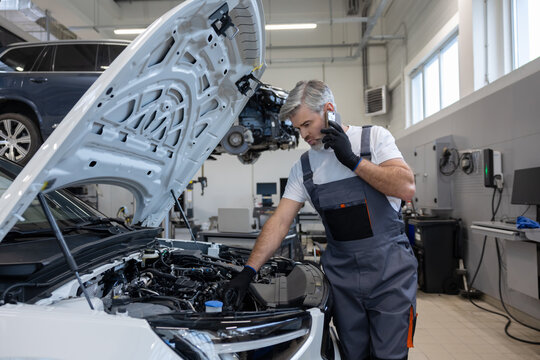 Engineer in workshop using mobile device for communication while working on vehicle engine fixing - Powered by Adobe