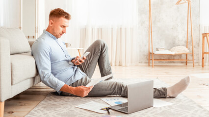 Young freelancer working from home, sitting on floor with pc, using smart phone and holding documents