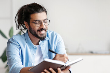 Smiling Western Freelancer Guy Taking Notes To Notepad At Workplace In Home Office, Planning Working Schedule, Filling Diary, Noting Business Ideas, Sitting At Desk With Laptop Computer, Copy Space