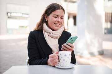 Young woman happily using her smartphone while enjoying a warm cup of coffee at a stylish cafe, immersed in her browsing experience and savoring every moment with her beverage