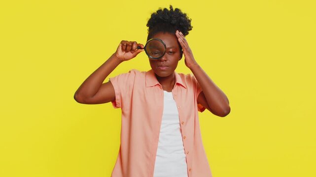 Investigator researcher scientist woman holding magnifying glass near face looking into camera with big zoomed funny eye, searching analyzing. Young African American girl isolated on yellow background