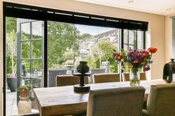 Bright and airy room featuring a wooden table, stylish decor, and large windows opening to a lush garden. A perfect blend of comfort and sophistication.