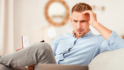 Planning And Project. Portrait of focused pensive man working from home, sitting on couch, holding notebook, looking at laptop. Guy watching webinar, thinking about remote work and his schedule