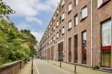 Fototapeta premium A contemporary urban building showcases its red brick facade along a tree-lined pathway, highlighting modern architecture and landscaping.