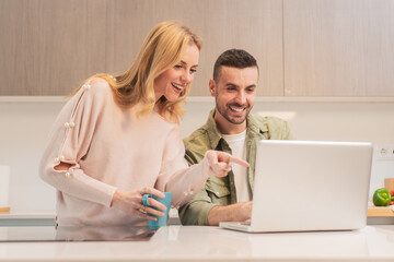 Joyful couple sharing exciting moments while engaging with a laptop in their cozy kitchen, filled with laughter and smiles during a delightful afternoon together, enjoying the time spent online