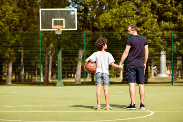 Obraz premium Back view full length portrait of mature man and boy standing on sports field with basket ball, holding hands