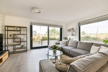 An elegant modern living room featuring minimalist furniture with a spacious layout, natural light, and greenery visible outside through large glass windows.