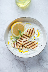 Bowl of greek tzatziki or cucumber yogurt sauce with grilled halloumi cheese, vertical shot on a white stone background, elevated view