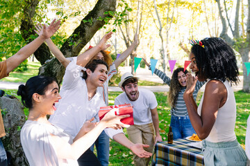 Excited friends surprising a young woman with a birthday gift at a cheerful outdoor party in a park
