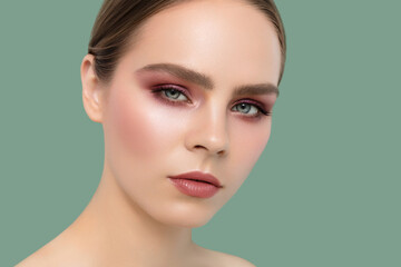 Confident and stylish, a young woman with slicked-back hair showcases bold pink eye makeup and glossy lips in a high-fashion beauty portrait against a green background..