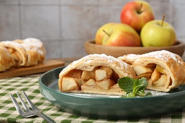 Pieces of tasty apple strudel with powdered sugar, mint and fork on table against grey tiled wall, closeup
