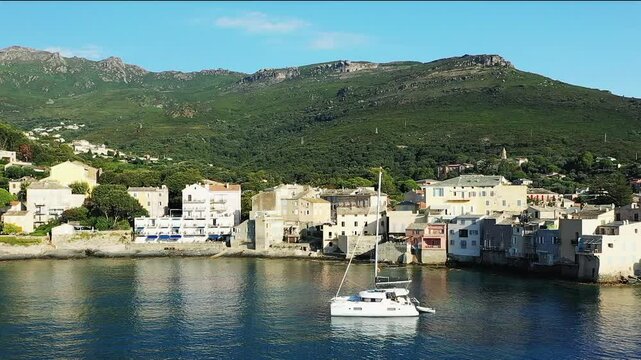 A boat sails in front of Erbalunga surrounded by dry and green mountains, in Europe, in France, in Corsica, towards Bastia, on the edge of the Mediterranean Sea, in summer, on a sunny day.&nbsp;
