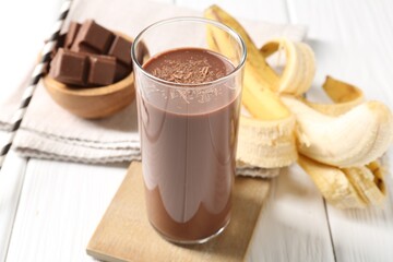 Tasty chocolate milk in glass, banana and pieces on white wooden table, closeup