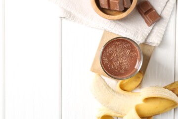 Tasty chocolate milk in glass, banana and pieces on white wooden table, flat lay