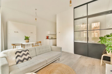 A stylish and modern minimalist living room featuring a cozy sofa, dining area, and kitchenette, complemented by natural light and greenery.