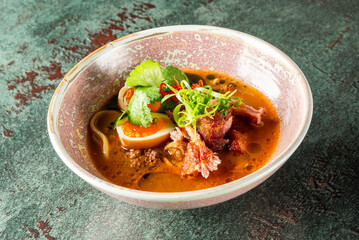 A rich and aromatic bowl of ramen featuring tender meats, a soft boiled egg, fresh herbs, and garnishes, served in an elegant pink bowl, perfect for warmth and flavor.
