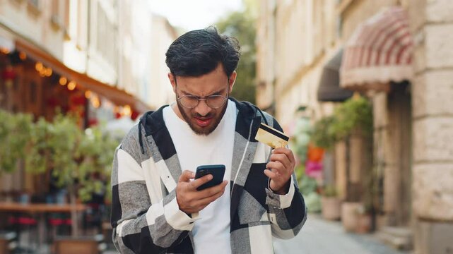 Shocked Hispanic man trying to pay online shopping with smartphone blocked credit card. Annoyed young Arabian tourist on street buying bank refuse problem unsuccessful payment lack of money balance