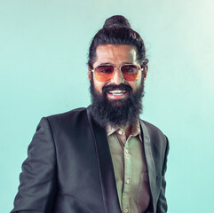 Fototapeta premium smiling man with long dark hair tied in a topknot, a full beard, and sunglasses. He is wearing a dark suit jacket over a light-colored collared shirt, standing against a pale teal background.